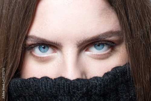 Beauty and fashion concept. Close-up woman portrait. Model with long dark hair and visible only blue eyes. Model hiding her mouth in sweater. Eyes is in camera focus