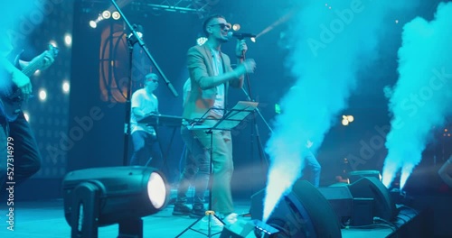 Band performs on stage at musical concert in spotlight and smoke. Vocalist actively moving and gesticulating. Slow motion