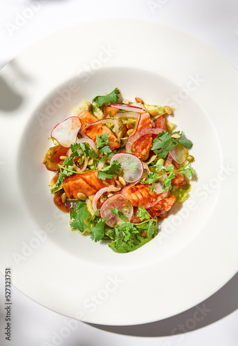 Elegant salad with baked salmon in sauce with vegetables on white table with harsh shadows. Salmon salad with radish on white background with shadows of leaves. Summer salad with roasted trout