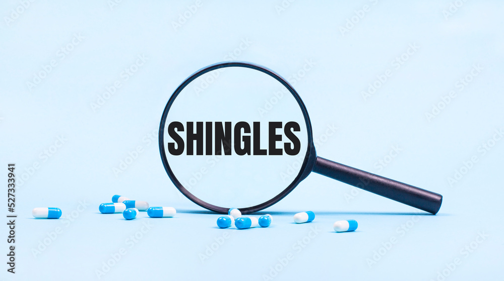On a blue background, white and blue capsules with pills and a black magnifying glass with the text SHINGLES. Medical concept.