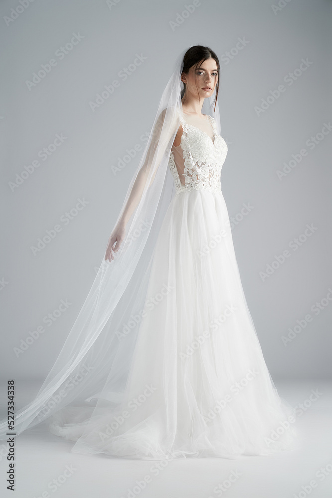 Portrait of a young beautiful bride in a long white wedding dress with lace and a long veil with a comb. The brunette girl is posing in a wedding dress on a grey background. Profile view.