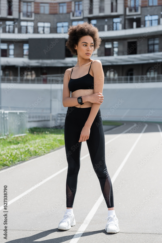 full length of african american woman in black sportswear and white sneakers standing on city stadium.