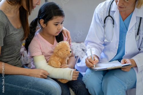 Pediatrician doctor examining little asian girl with a broken arm wearing a cast at hospital photo