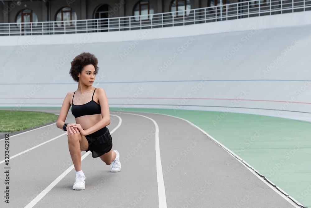 Young african american sportswoman doing lunges on running track of stadium.
