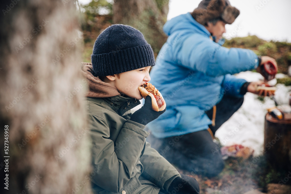 Boy in warm clothing eating hot dog while father preparing food Boy in warm clothing eating hot dog while father preparing food
