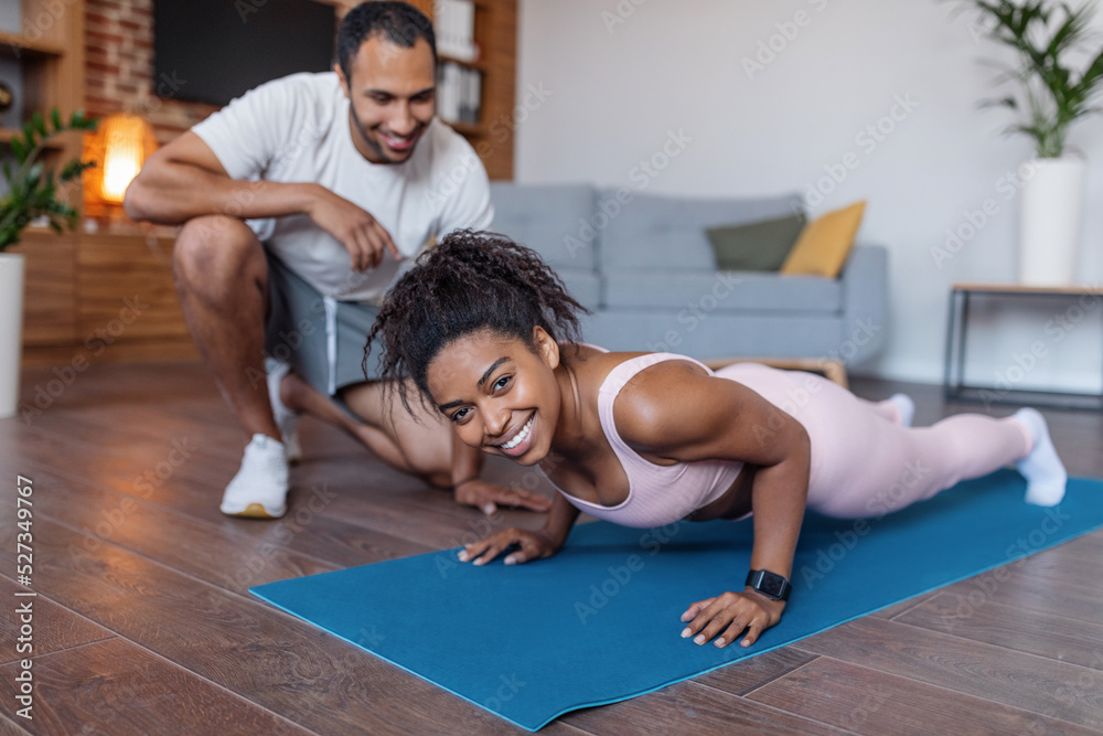 Naklejka premium Cheerful millennial african american man in sportswear helping to wife doing push ups in living room interior
