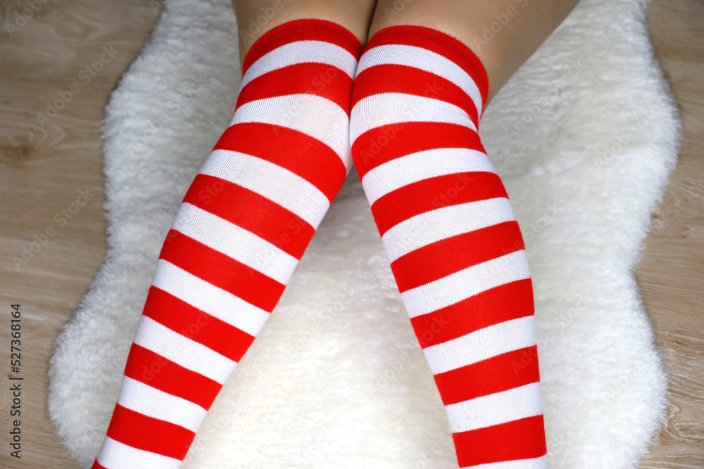 Fototapeta premium Legs of girl in Christmas knee socks sitting on white fur rug on wooden floor. Female outfit for New Year celebration