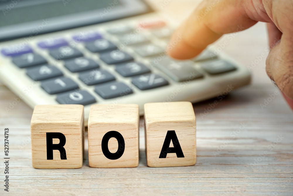 ROA - acronym from wooden blocks with letters. business concept finger background with blur calculator.  ROA - short for Return On Assets                 