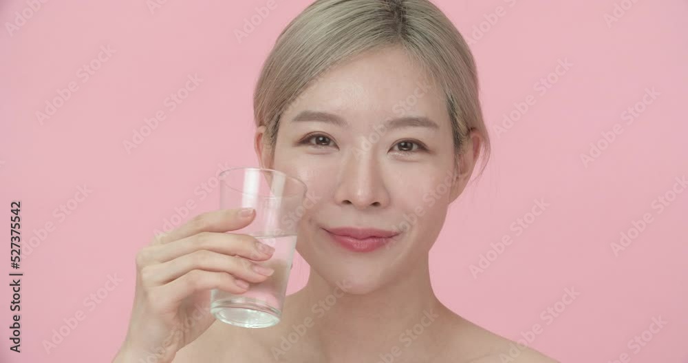 Studio portrait of a young, beautiful Asian woman with blond hair holding glass, then drinking water and smiling at the camera in pink background. Blogger with beauty, healthy or fashion concept. Studio portrait of a young, beautiful Asian woman with blond hair holding glass, then drinking water and smiling at the camera in pink background. Blogger with beauty, healthy or fashion concept.