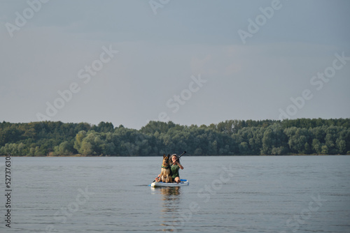 Stand up paddle with pet. Concept animals actively spend time with owner. Young Caucasian woman with dreadlocks sits on inflatable board in lake and paddles with German Shepherd in a life jacket.