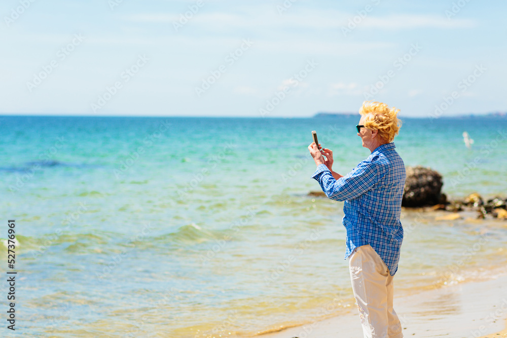 Elderly woman standing on the beach by the sea and taking photo of the sea and seagulls on mobile phone. Active seniors concept