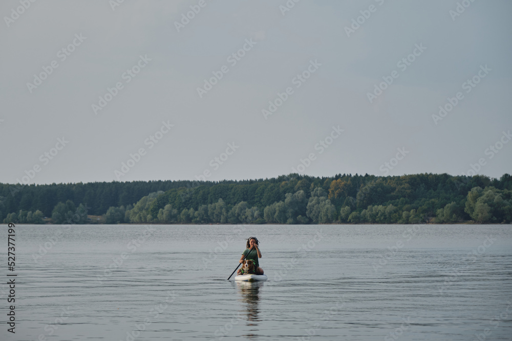 Caucasian woman with dreadlocks sitting on board in lake and rowing with oar. Australian Shepherd in life jacket. Stand up paddle with pet. Concept animals actively spend time with owner.