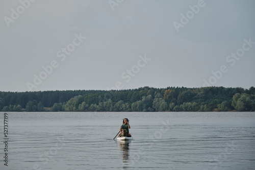 Caucasian woman with dreadlocks sitting on board in lake and rowing with oar. Australian Shepherd in life jacket. Stand up paddle with pet. Concept animals actively spend time with owner.
