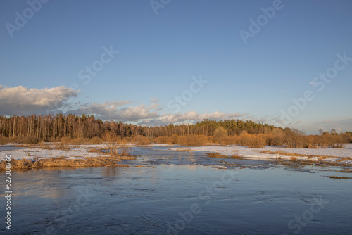 March sunny evening by the river. Blue sky over the horizon. A picturesque landscape, early spring, a river with snow-covered banks, dry grass and bushes. The first thaws, the snow is melting