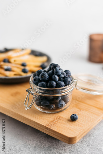 a jar of blueberries on a wooden board, next to pancakes. light background