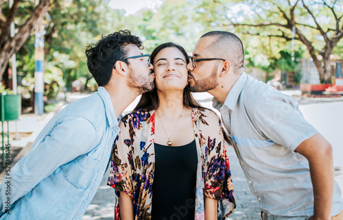 Love triangle concept. Polygamy concept. Two men kissing a girl cheek. Portrait of two guys kissing a girl cheek. Two young men kissing a woman cheek outdoor photo