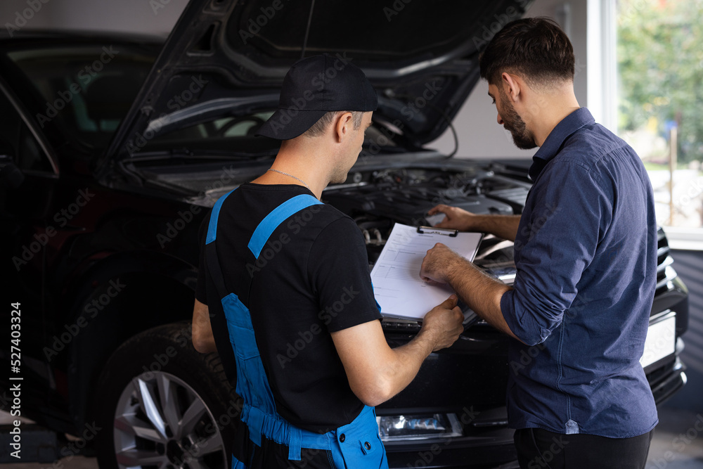 Customer and mechanic discuss upcoming car repair. Auto service, repair, deal and people concept. Mechanic and car owner signing paper at workshop.