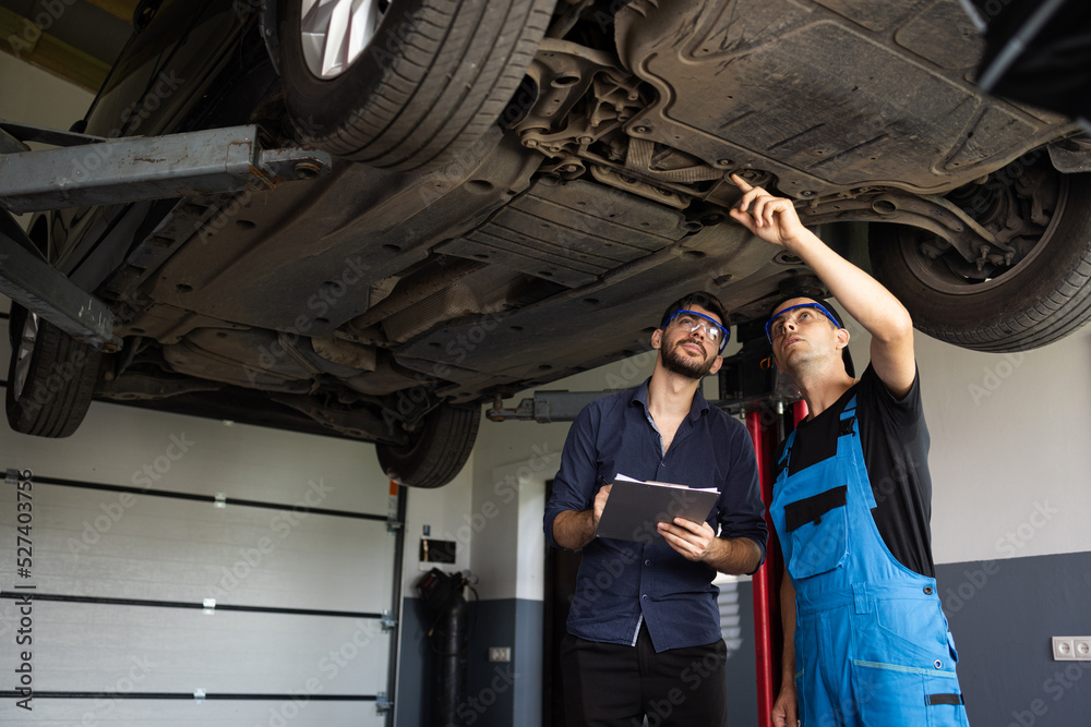 Auto Service. Car Service Employees Inspect the Bottom and Skid Plates of the Car. Manager Checks Data on a Notebook and Explains the Breakdown to a Mechanic. Modern Workshop