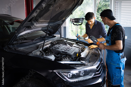 Car service employees inspect car's engine bay with a LED lamp. Manager checks tasts on tablet computer and explains an Engine breakdown to an male mechanic. Auto service