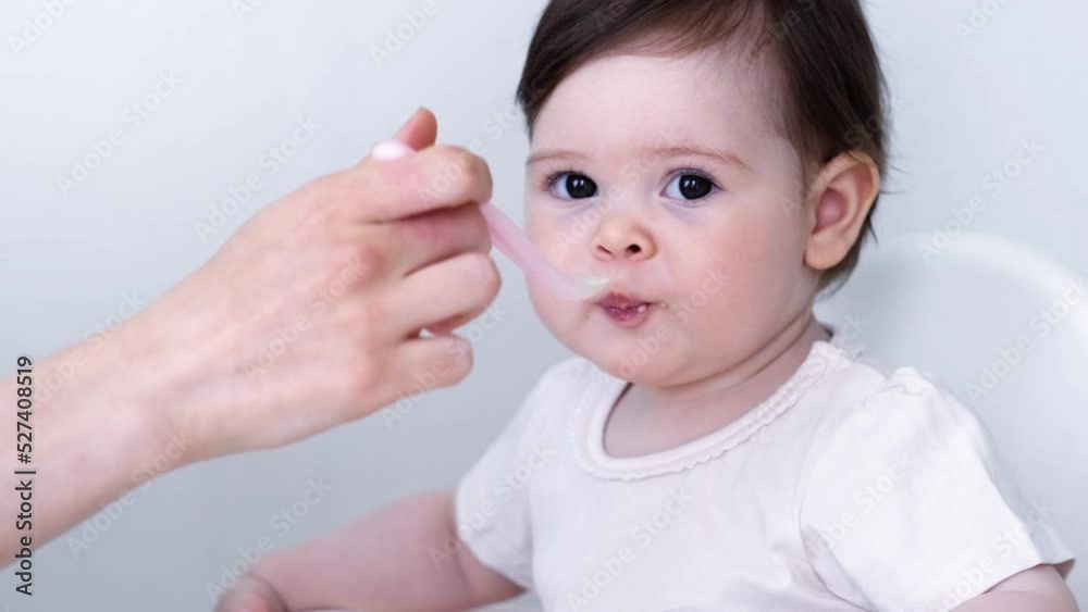 Baby girl eating blend mashed food sitting, on high chair, mother feeding child, hand with spoon for vegetable lunch, baby weaning, first solid food for young kid. Baby girl eating blend mashed food sitting, on high chair, mother feeding child, hand with spoon for vegetable lunch, baby weaning, first solid food for young kid.