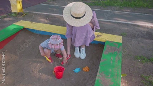 Mom in a purple dress and a straw hat plays with her 2-year-old daughter in the sandbox on the playground