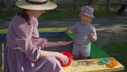 Mom plays with a 2-year-old baby in the sandbox with plastic toys with a shovel and a bucket