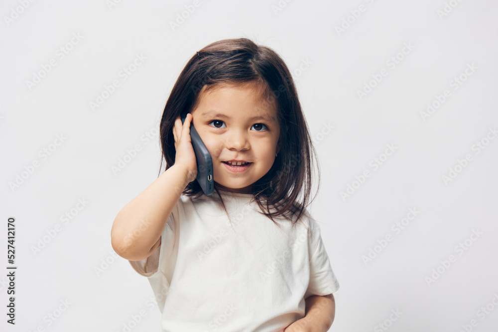 a little girl of preschool age, sitting on a white background in a white T-shirt, enthusiastically talking on the phone, turning half-side to the camera. The topic of the conversation on the phone a little girl of preschool age, sitting on a white background in a white T-shirt, enthusiastically talking on the phone, turning half-side to the camera. The topic of the conversation on the phone