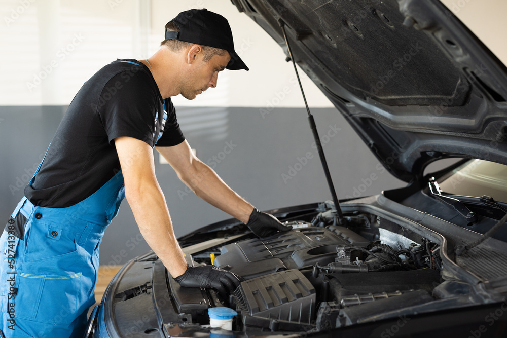 Mechanic man open a car hood and check up the engine. Car mechanic noting repair parts during open car hood engine repair at garage. Overheating of a car engine. Motor with open hood Mechanic man open a car hood and check up the engine. Car mechanic noting repair parts during open car hood engine repair at garage. Overheating of a car engine. Motor with open hood