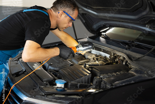 Caucasian male mechanic in blue uniform checking motor in auto service with light. Young man engineer fixing engine. Maintaining concept. Repairment work of technician