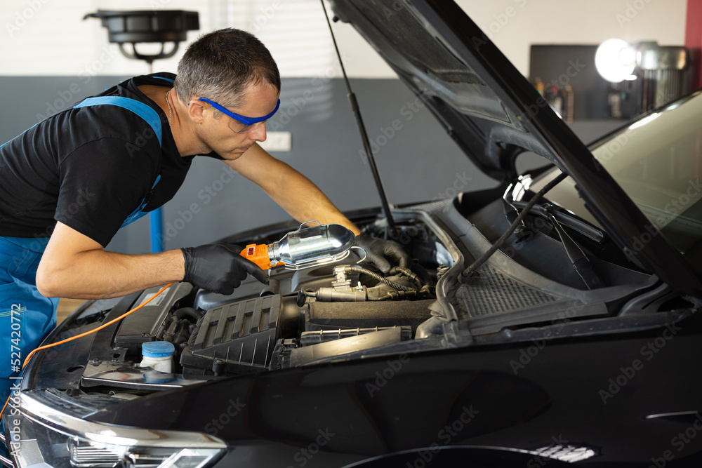 Young caucasian man in blue overalls and safety glasses inspects engine with flashlight. Male car mechanic at work in spacious repair shop. Modern workshop Young caucasian man in blue overalls and safety glasses inspects engine with flashlight. Male car mechanic at work in spacious repair shop. Modern workshop