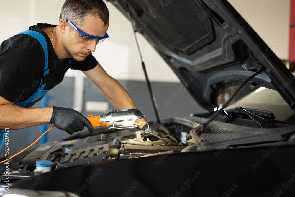 Caucasian Mechanic in Blue Overalls is Working on a Car in a Car Service. Repairman in Safety Glasses is Working on an Usual Car Maintenance. He Hangs Led Lamp. Modern Workshop Caucasian Mechanic in Blue Overalls is Working on a Car in a Car Service. Repairman in Safety Glasses is Working on an Usual Car Maintenance. He Hangs Led Lamp. Modern Workshop