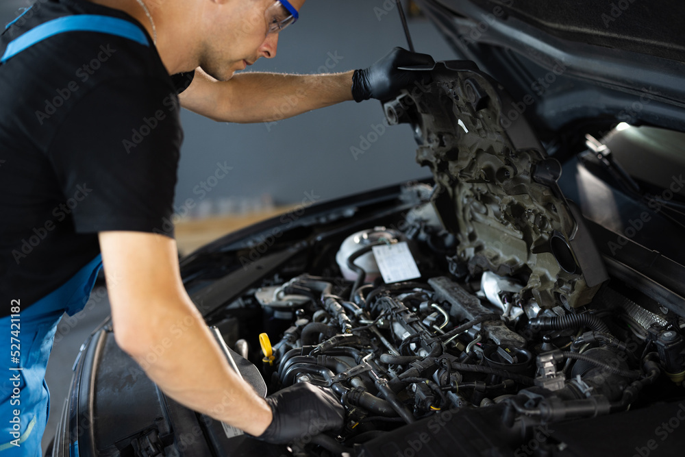 Car mechanic noting repair parts during open car hood engine repair at garage. Unrecognizable mechanic man open a car hood and check up the engine. Overheating of a car engine. Motor with open hood Car mechanic noting repair parts during open car hood engine repair at garage. Unrecognizable mechanic man open a car hood and check up the engine. Overheating of a car engine. Motor with open hood