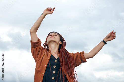 Pretty woman in boho clothes with red dreadlocks is dancing on the rooftops against the background of a gray sky. Hope. Autumn color and mood. Fresh air.