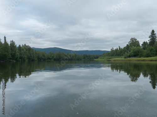 View of wide calm Tarra river from boat at Kvikkjokk, with forest and grass shore and green hills. Northern landscape in Swedish Lapland. End of Padjelantaleden hiking trail. Cloudy summer day