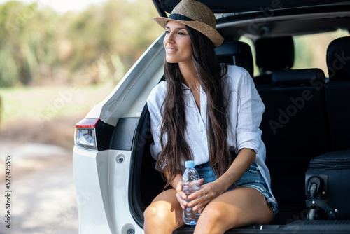 Beutiful woman sitting in the trunk of the car while drinking water in the forest.