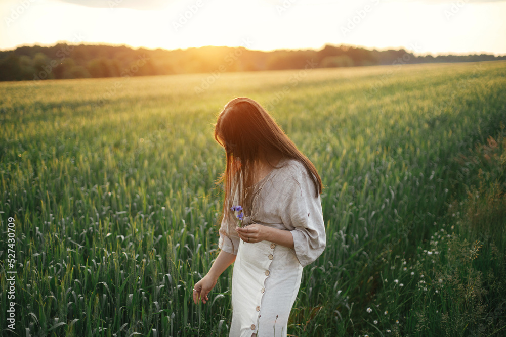 Woman relaxing in wheat field in warm sunset light. Stylish young female in rustic dress holding wildflowers in hands in evening summer countryside. Tranquil atmospheric moment