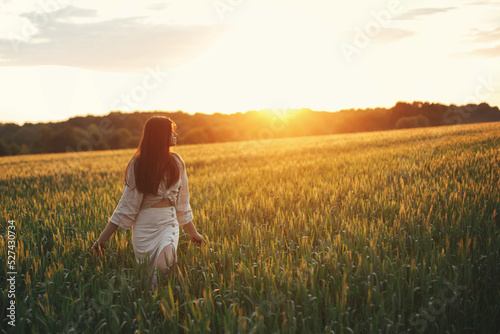 Woman walking in wheat field in warm sunset light. Stylish young female in rustic dress holding wildflowers in hands and relaxing in evening summer countryside. Tranquil atmospheric moment