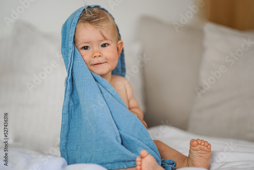 mega cute naked almost one year old blond baby boy sitting & laughing at home on a cozy bed after bathing and playing with a blue muslin fabric burp cloth while making nonsense jokes