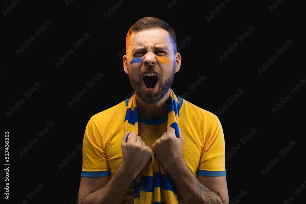 Portrait of excited young soccer supportred man in yellow-blue t-shirt and scarf and painted face, cheering for his favourite team Portrait of excited young soccer supportred man in yellow-blue t-shirt and scarf and painted face, cheering for his favourite team