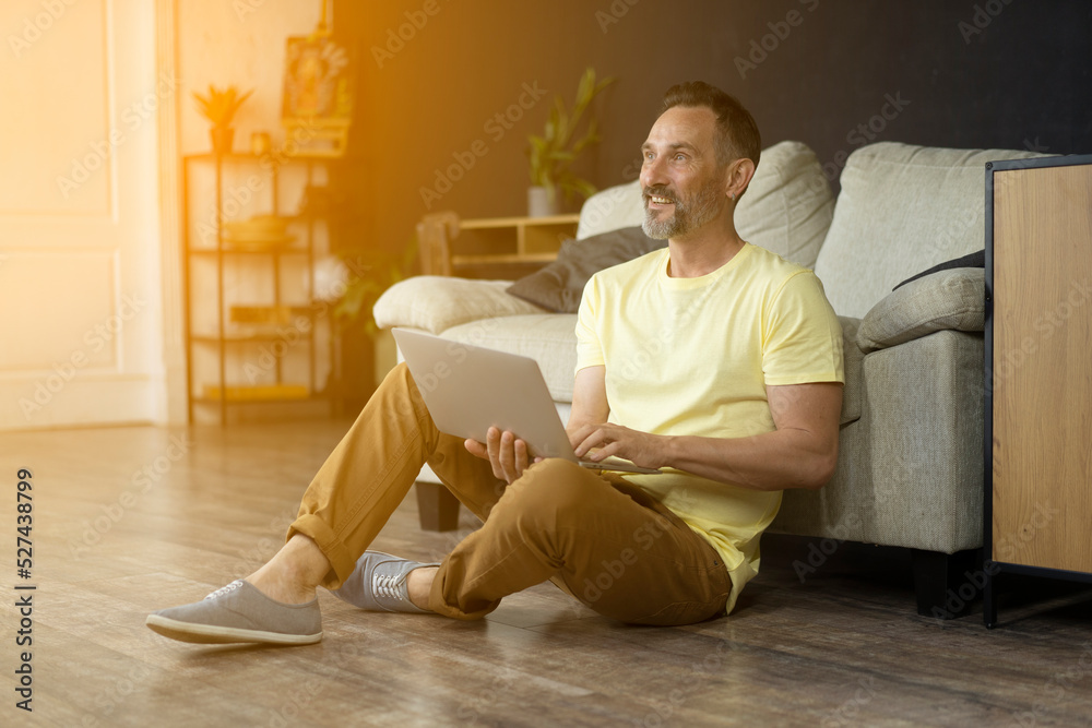 Smiling mature man on floor indoor sitting near of sofa in living room working on laptop compunter Handsome middle-age bearded man looking into sunshine light at home,