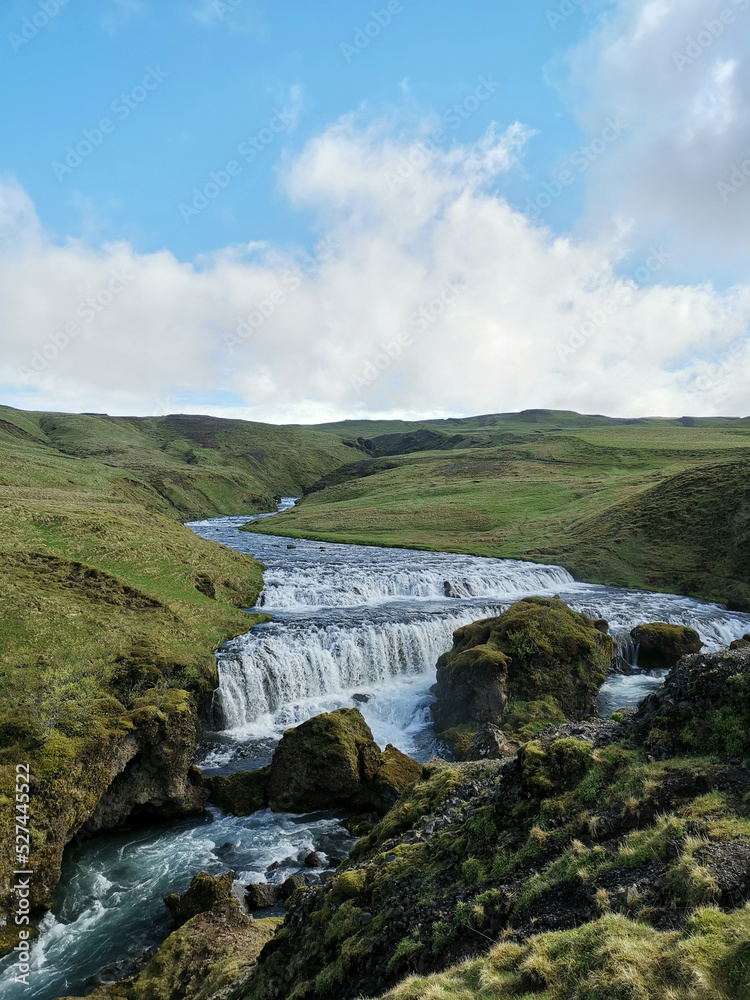 Fototapeta premium River going through mountains and green grass with water flowing stone waterfall with blue sky and white cloud vertical idyllic nature
