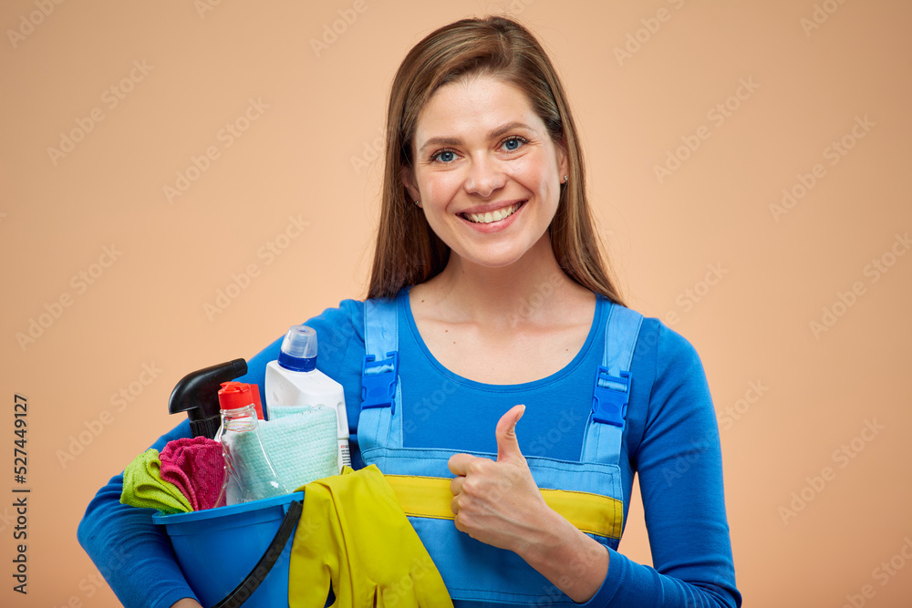 Smiling woman in blue overalls holding cleaning detergent in bucket. Thumb up. isolated on brown beige background. Smiling woman in blue overalls holding cleaning detergent in bucket. Thumb up. isolated on brown beige background.