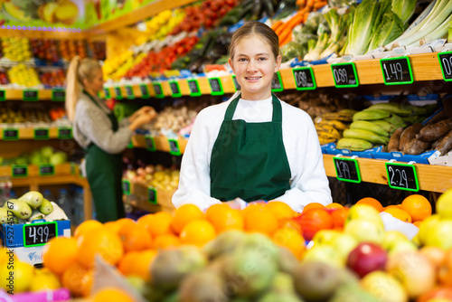 Portrait of smiling young girl wearing apron standing in fruit and vegetable store. First job concept..