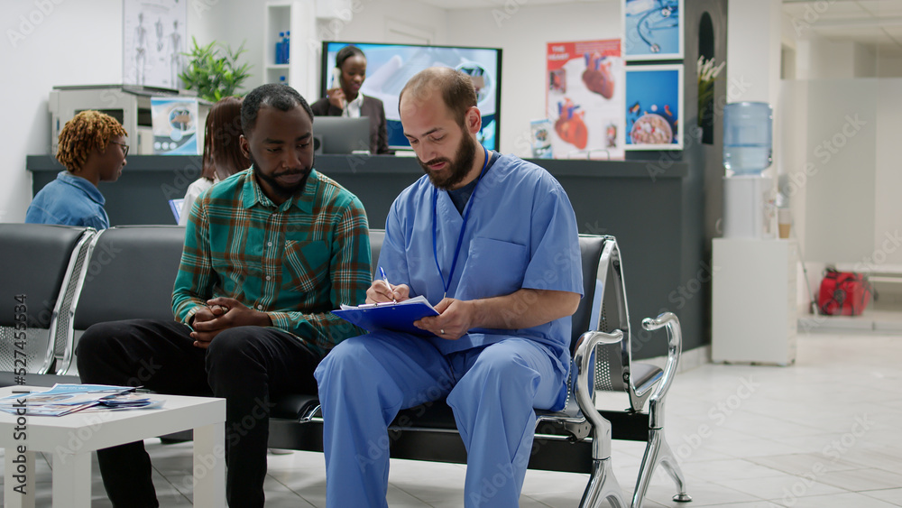 Male nurse talking to african american man in waiting room lobby, doing checkup consultation in hospital reception area. People discussing about healthcare recovery at examination.