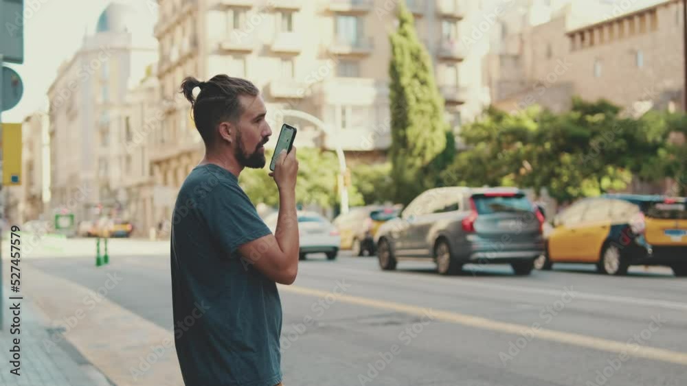 Young man with beard uses cellphone while standing on crosswalk, sends voice message Young man with beard uses cellphone while standing on crosswalk, sends voice message