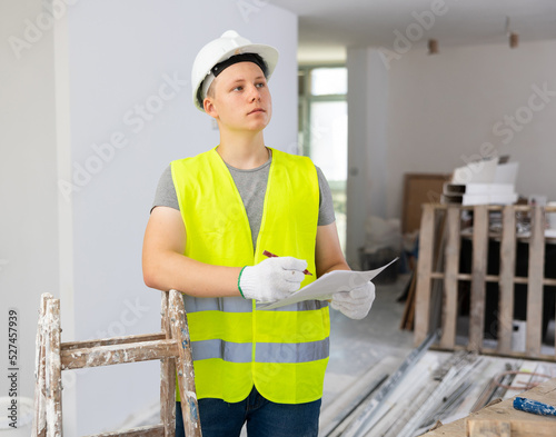 Fifteen-year-old worker in a yellow vest, working on a construction site indoors, makes important notes on paper, studying ..the house repair plan photo