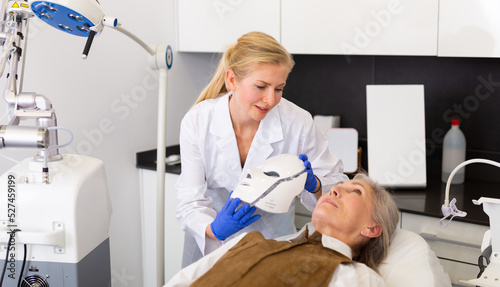 Young woman cosmetologist putting spectrum mask on senior woman s face.