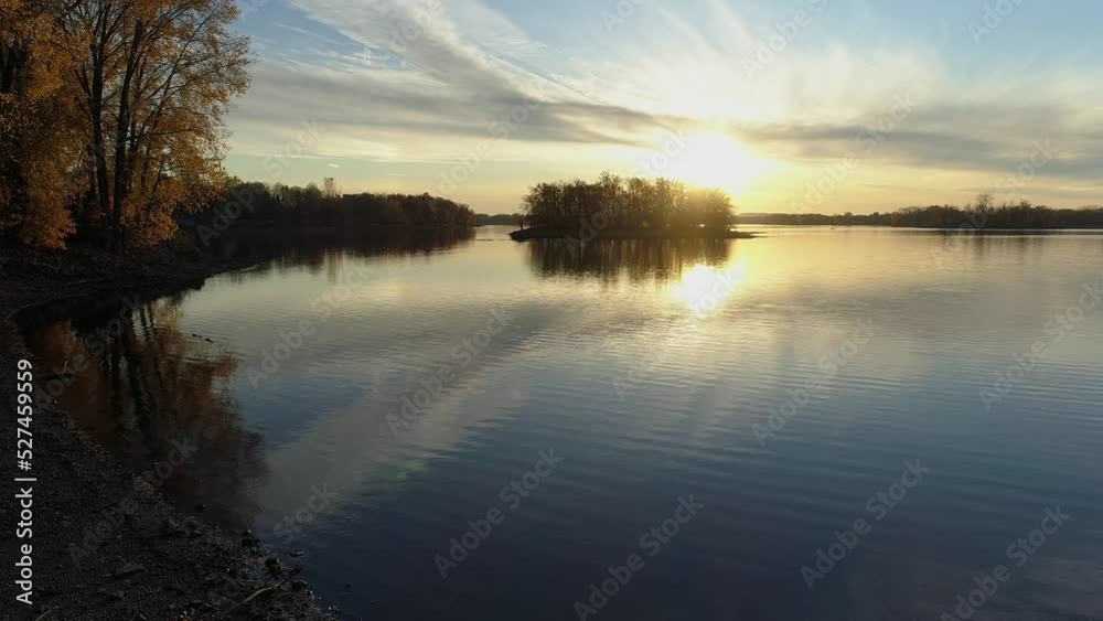 Aerial drone video moving backward over the shore, at low angle view, keeping the sunset and colorful trees in view Aerial drone video moving backward over the shore, at low angle view, keeping the sunset and colorful trees in view