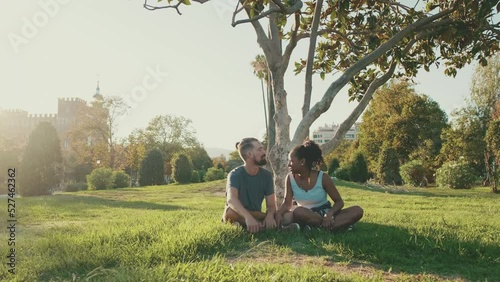 Happy smiling interracial couple talking while sitting on the grass in the park. Backlight