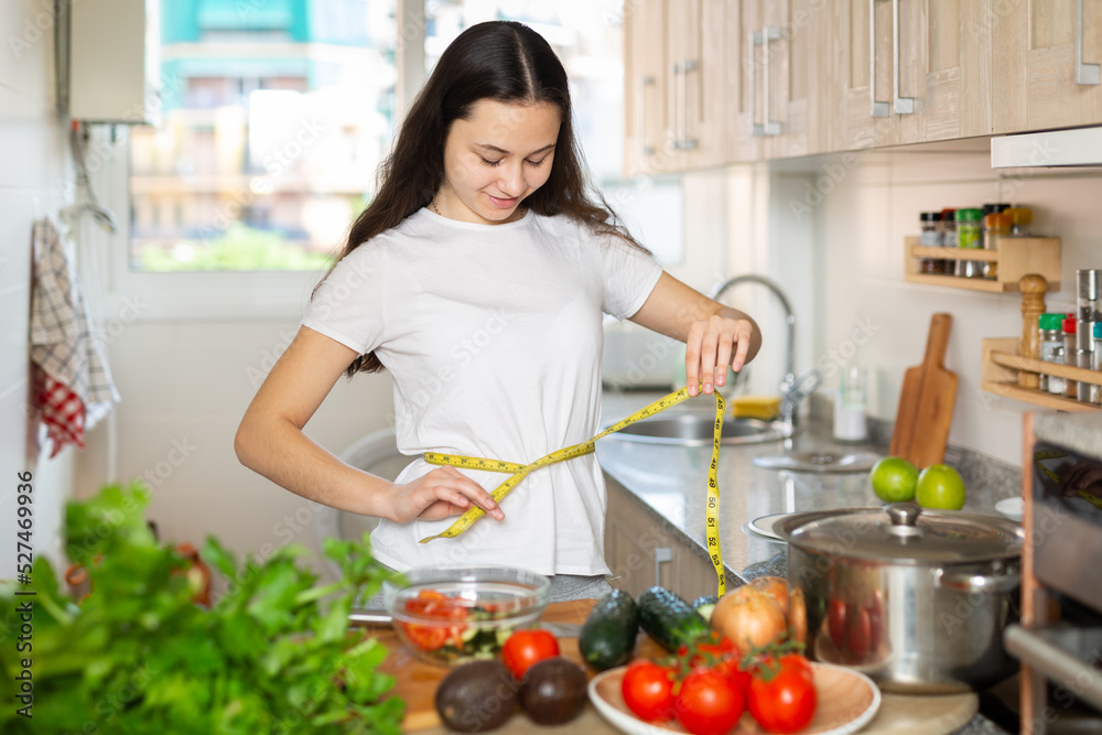 Young cheerful woman taking measurements of her body using tape measure while preparing salad at kitchen